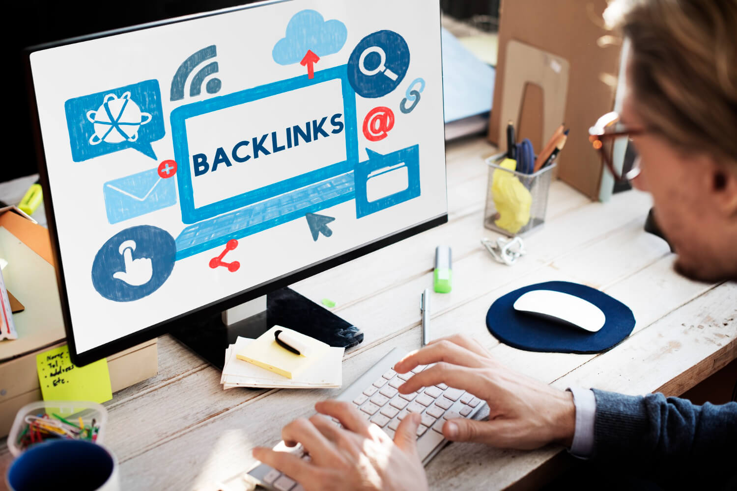 Un hombre con barba y gafas trabaja en un ordenador de escritorio. En la pantalla del monitor, se muestra una ilustración digital con la palabra 'BACKLINKS' destacada en el centro, dentro de un recuadro que simula una pantalla de ordenador. Alrededor de este recuadro central, hay diversos iconos relacionados con el marketing digital y la conexión en línea, como un símbolo de Wi-Fi, una nube con una flecha hacia arriba, una lupa, el símbolo de arroba (@), un sobre, un pulgar hacia arriba y flechas de compartir en redes sociales. El hombre está tecleando en un teclado blanco, con una mano cerca del ratón blanco sobre una alfombrilla azul. El escritorio está desordenado con notas adhesivas, bolígrafos y otros objetos de oficina. La imagen ilustra visualmente el concepto de backlinks y la importancia de las conexiones en línea para el SEO Offpage.