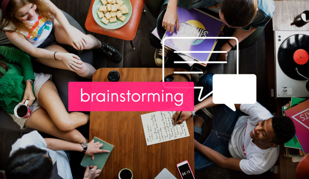 Vista aérea de un grupo de jóvenes diversos (hombres y mujeres) sentados alrededor de una mesa de madera, inmersos en una sesión de 'Brainstorming y Listado Inicial' de ideas. Un gran cuadro de texto rosa brillante superpuesto en el centro de la imagen contiene la palabra 'brainstorming' en blanco. Se observa una mano escribiendo en un cuaderno con apuntes, mientras otros participantes sostienen tazas de café, un smartphone, y una tableta. Un tocadiscos vintage y una pila de discos de vinilo añaden un toque creativo al ambiente relajado. Esta imagen ilustra visualmente el proceso creativo de generación de ideas y palabras clave, fundamental para el contenido y el SEO en un blog. Representa a emprendedores digitales que buscan entender cómo funciona el proceso de inicio de un proyecto y están dispuestos a estudiar a profundidad cómo generar ideas de contenido y palabras clave relevantes para el posicionamiento a largo plazo de su sitio.