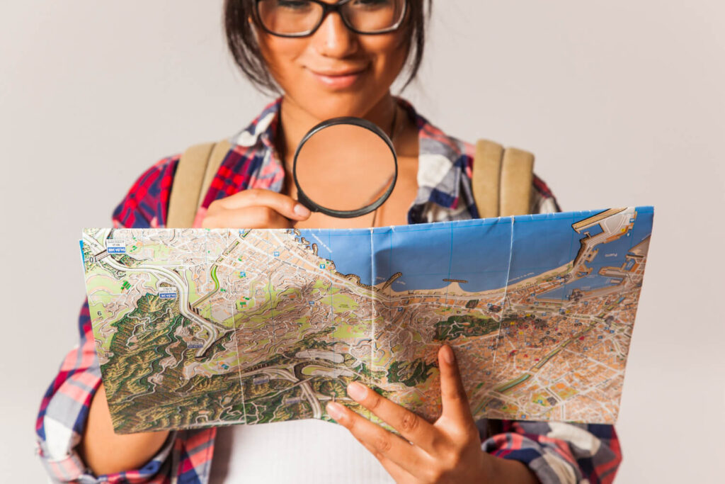 Una joven emprendedora digital con gafas y una mochila, vestida con una camisa de cuadros, sostiene un mapa abierto y lo examina con una lupa. Su expresión es de concentración y curiosidad, como si estuviera buscando un camino o planificando una ruta. El mapa muestra detalles geográficos y urbanos. Esta imagen representa visualmente la fase inicial de planificación y exploración necesaria para ¿Cómo empezar con SEO? Pasos clave.