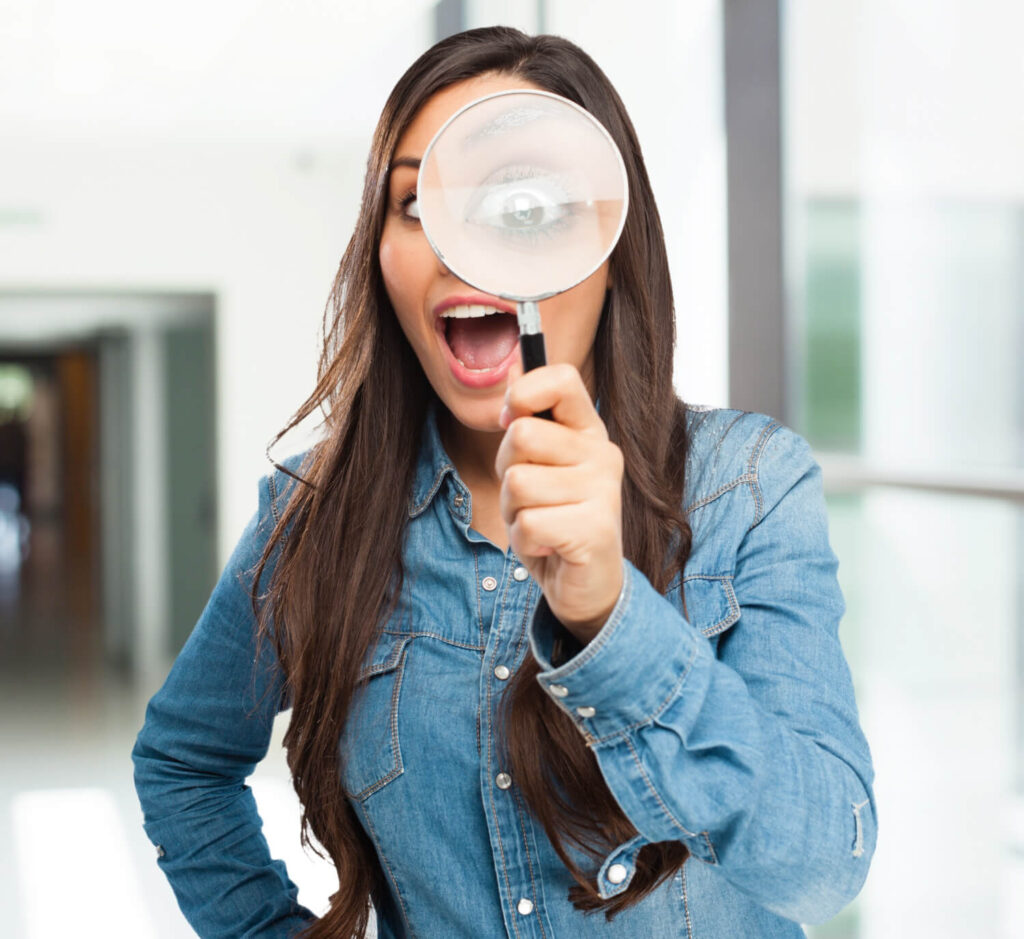 Una mujer joven con cabello largo y castaño, vestida con una camisa de mezclilla azul, mira a través de una lupa grande que sostiene frente a su ojo derecho. Su boca está abierta en una expresión de sorpresa o asombro, y su ojo a través de la lupa se ve ampliado. El fondo es un interior borroso que sugiere un pasillo o una oficina. Esta imagen ilustra visualmente la acción de una búsqueda o investigación detallada, que es esencial para comprender 'La Intención de Búsqueda: El Núcleo del Contenido' dentro del blog 'Contenido y Palabras Clave'. Representa a los emprendedores digitales que, con una mirada a largo plazo, están dispuestos a estudiar a profundidad cómo funciona el SEO, específicamente la importancia de identificar qué hay detrás de las palabras clave para crear contenido de alta calidad que resuene con la intención de búsqueda de sus usuarios.
