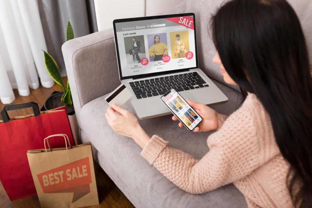 Una mujer con cabello oscuro y un suéter rosa claro está sentada en un sofá gris, realizando compras en línea desde un portátil y un teléfono móvil. El portátil muestra una tienda de moda en línea con la etiqueta 'SALE' y descuentos. La mujer sostiene una tarjeta de crédito en una mano y mira la pantalla de su teléfono móvil con la otra, donde también se visualizan productos de moda. A la izquierda, se observan bolsas de compra, una de ellas con la etiqueta 'BEST SALE'. Esta imagen ilustra la actividad de compra en línea, un comportamiento donde la visibilidad a través del ¿Por qué el SEO es clave en el marketing digital? se vuelve fundamental para los emprendedores digitales que buscan entender cómo atraer clientes a largo plazo. El SEO permite que sus negocios sean encontrados por usuarios que buscan productos como los mostrados, comprendiendo así su funcionamiento y la importancia en el panorama del marketing digital.