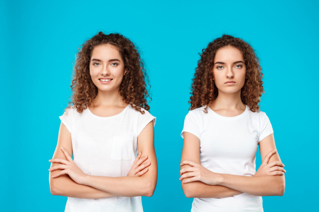 Dos mujeres jóvenes con cabello rizado y castaño, vestidas con camisetas blancas idénticas, posan una al lado de la otra sobre un fondo azul brillante. La mujer de la izquierda sonríe amistosamente con los brazos cruzados, mientras que la mujer de la derecha tiene una expresión más seria o neutra, también con los brazos cruzados. La similitud casi perfecta entre ambas mujeres sugiere una representación visual de la duplicidad o repetición. Esta imagen es ideal para ilustrar el subcapítulo 'Google penaliza automáticamente el contenido duplicado' dentro del blog 'Mitos sobre el posicionamiento en Google', ayudando a los emprendedores digitales a entender cómo funciona realmente el contenido duplicado y por qué no siempre resulta en una penalización automática por parte de Google, desmintiendo un mito común en el mundo del SEO y el posicionamiento.