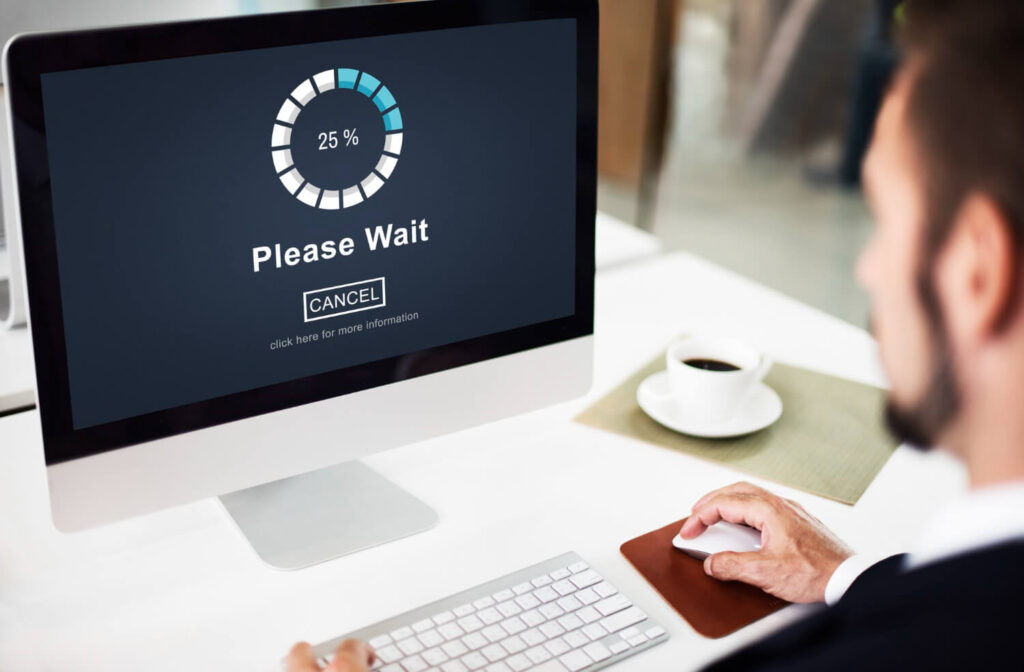 Un primer plano de un ordenador de escritorio mostrando una pantalla con un mensaje de 'Please Wait' y una barra de carga circular que indica un progreso del 25%. Debajo del mensaje, hay un botón de 'Cancel' y un texto más pequeño que dice 'click here for more information'. Una mano de un hombre se ve utilizando un ratón blanco sobre una alfombrilla marrón a la derecha del teclado blanco. A la derecha del monitor, hay una taza blanca con café sobre un plato y un mantel individual verde claro. Esta imagen ilustra visualmente una situación de espera durante la carga de recursos, lo cual es directamente relevante para el concepto de Optimización de Imágenes y Recursos Multimedia para mejorar la velocidad de carga de un sitio web.