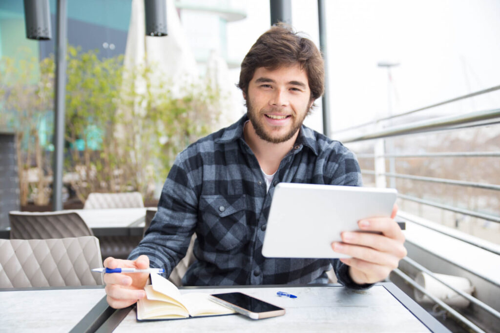 Un joven emprendedor digital con barba incipiente y cabello castaño está sentado en una mesa al aire libre, sonriendo mientras sostiene una tableta plateada en su mano derecha. En la mesa, frente a él, hay un cuaderno abierto, un bolígrafo azul y un smartphone negro. El fondo muestra un entorno exterior con vegetación y elementos arquitectónicos modernos. Esta imagen ilustra visualmente a un individuo aprendiendo y trabajando con herramientas digitales, lo cual es relevante para el subcapítulo '¿Por qué aprender SEO desde 0?' dentro del blog 'SEO desde Cero: Cómo Google Decide Quién Gana el Juego del Posicionamiento'. Representa a los emprendedores digitales que están dispuestos a estudiar a profundidad el SEO para entender cómo Google decide el juego del posicionamiento y así aplicarlo a largo plazo en sus estrategias digitales.